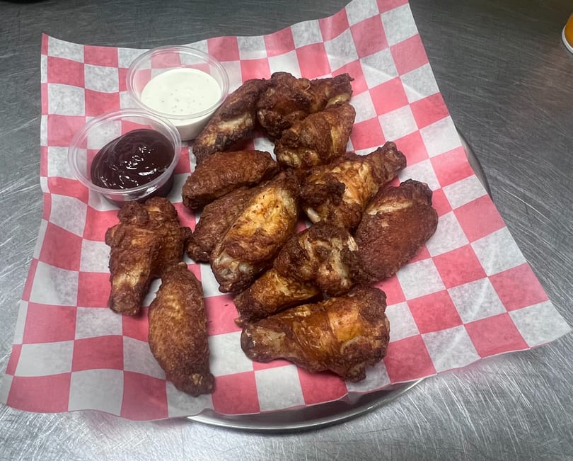 Crispy fried chicken wings with barbecue and ranch dipping sauces on red and white checkered paper.