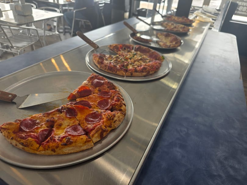 Multiple pizzas with various toppings, including pepperoni, on metal trays in a restaurant setting.