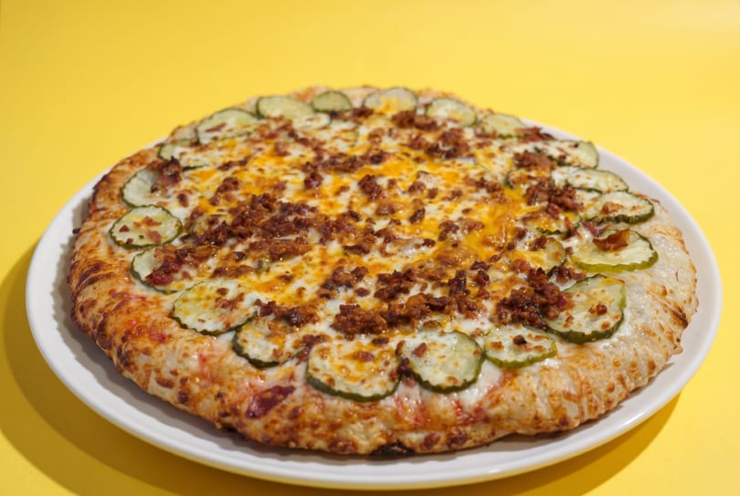A whole pizza topped with melted cheese, crumbled bacon, and pickle slices on a white plate.