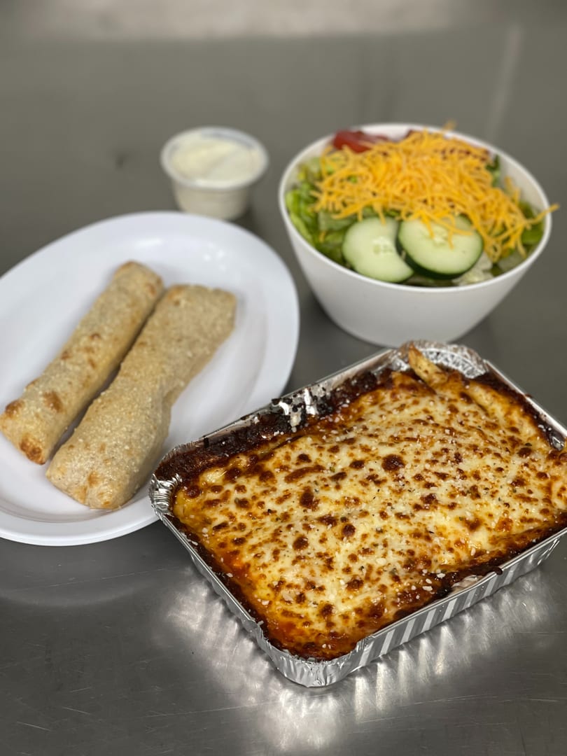 Baked lasagna in a foil pan, breadsticks, and a side salad with dressing on a metal surface.