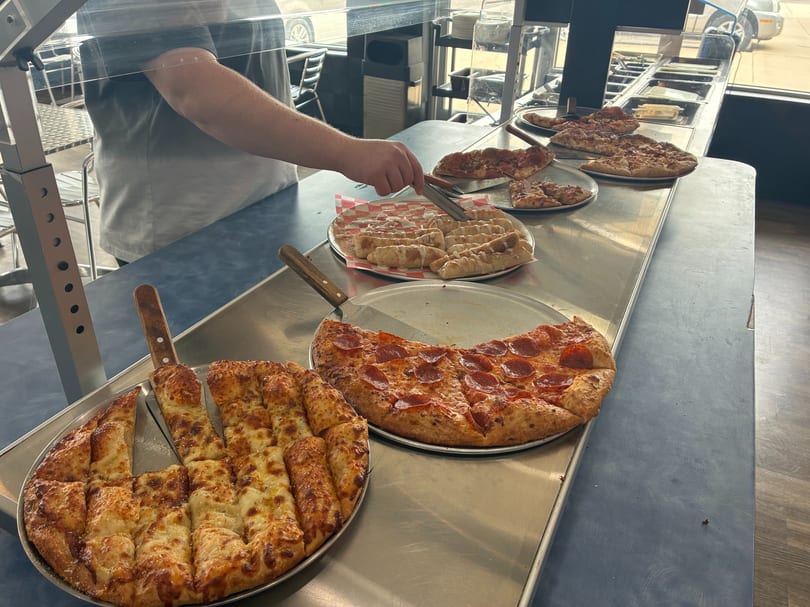 A person serving cinnamon rolls from a buffet line with various pizzas and breadsticks.
